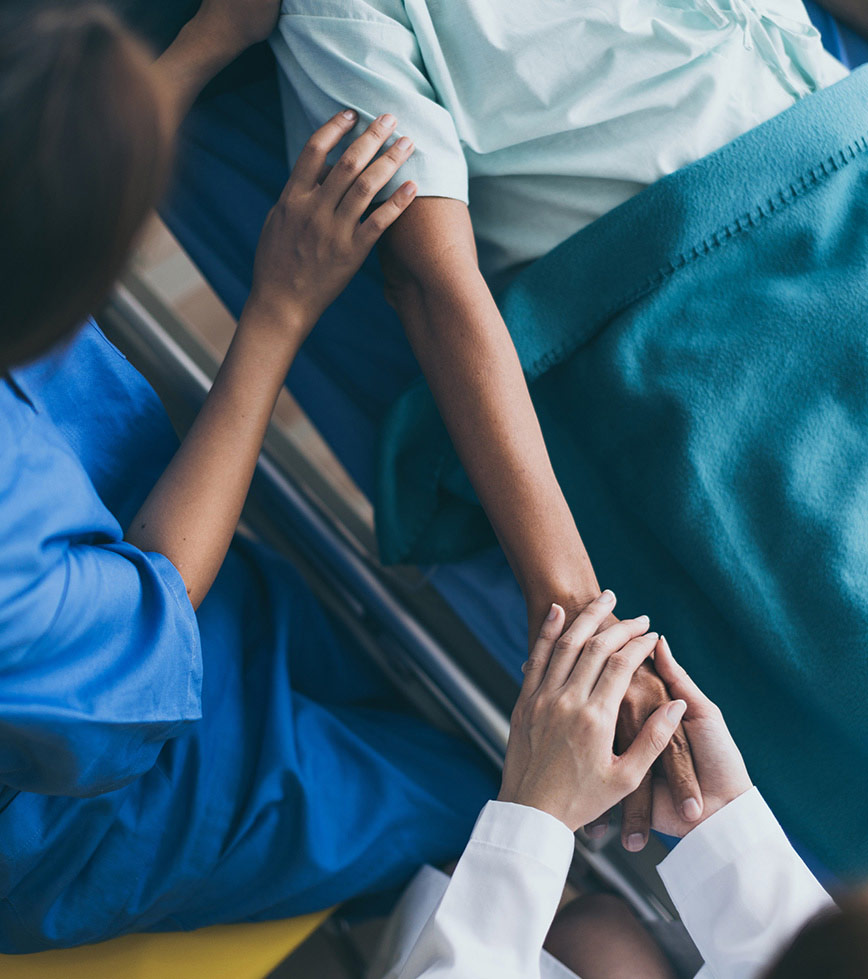 person in hospital bed holding someone's hand while medical person touches their shoulder