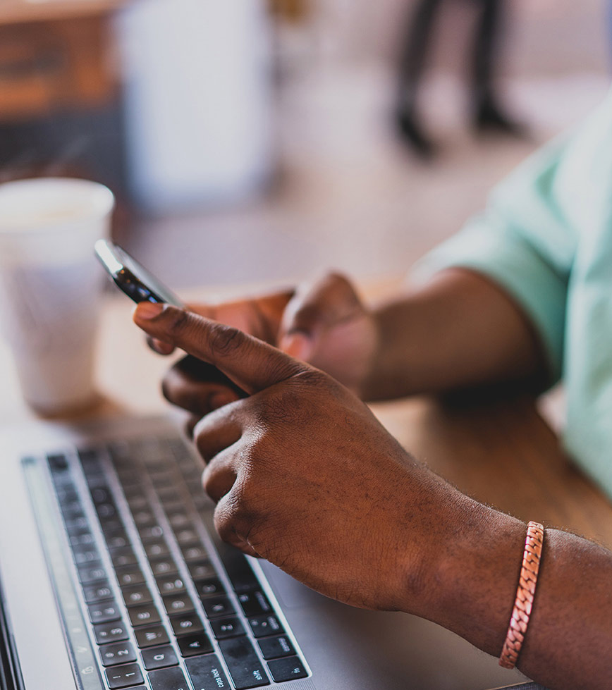 hands holding smartphone over desk with computer in coffee shop