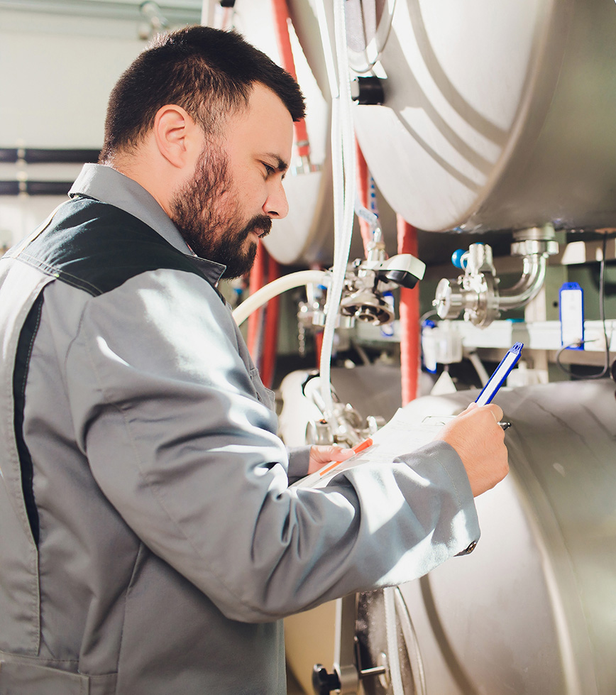 Man in long sleeve grey work shirt writing on a clipboard while inspecting metal barrels
