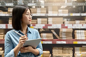 Woman with glasses holding tablet and pen looking left with inventory of boxes behind her